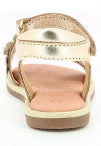 Sandale strappy en métal doré avec semelle plate, semelle intérieure en cuir brun, et accents texturés. Dos ouvert avec détail floral sur la sangle.