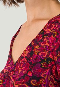 Femme montrant le haut de sa poitrine et son cou, vêtue d'un vêtement à col en V profond avec un motif floral paisley vibrant en rouge, rose, orange et noir.