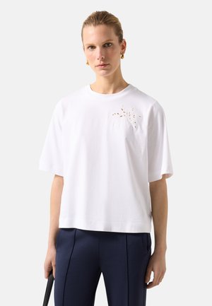 Kvinde iført en hvid kortærmet skjorte med et diskret bladudskåret design på brystet, marineblå bukser og gyldne creoløreringe.