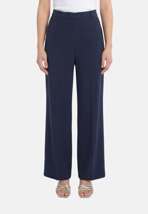 Persona che indossa pantaloni larghi blu navy, top bianco e sandali beige con cinturini, in piedi contro uno sfondo semplice.