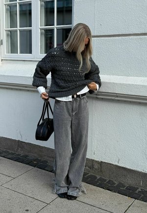 Hosszú szőke hajú nő sötét kötött pulóvert igazít, laza szürke farmert visel, fekete kézitáskát tart, egy ablakos fehér épület mellett áll.