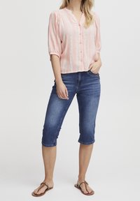 Camicia abbottonata rosa chiaro con strisce verticali, maniche a palloncino, abbinata a capri in denim blu di lunghezza al ginocchio e sandali marroni.