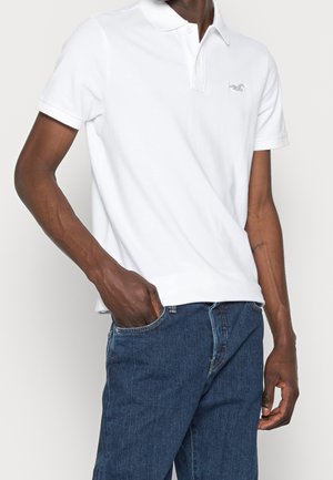 Person trägt ein weißes kurzärmeliges Poloshirt mit kleinem Logo auf der Brust und blaue Jeans, eine Hand in der Tasche.