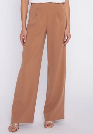 Kobieta w wysokich, szerokich spodniach w kolorze wielbłądzim, zestawionych ze złotymi sandałami na paski, stoi na tle jednolitego tła.