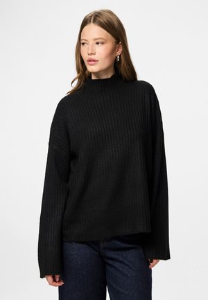 Femme aux longs cheveux bruns portant des boucles d'oreilles en argent, un pull à côtes noir et un jean bleu foncé, regardant vers sa gauche.