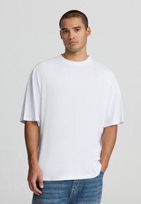 Biała, oversize'owa koszulka wykonana z bawełny, z krótkimi rękawami, okrągłym dekoltem i luźnym krojem, w zestawie z jasnoniebieskimi dżinsami.