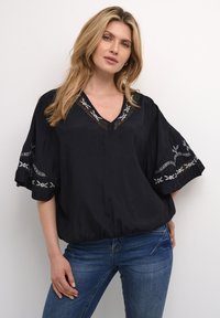 Blusa nera con scollatura a V e accenti floreali ricamati sulle maniche. Design morbido e fluido, texture delicata e fondo arricciato. Indossata con jeans blu.