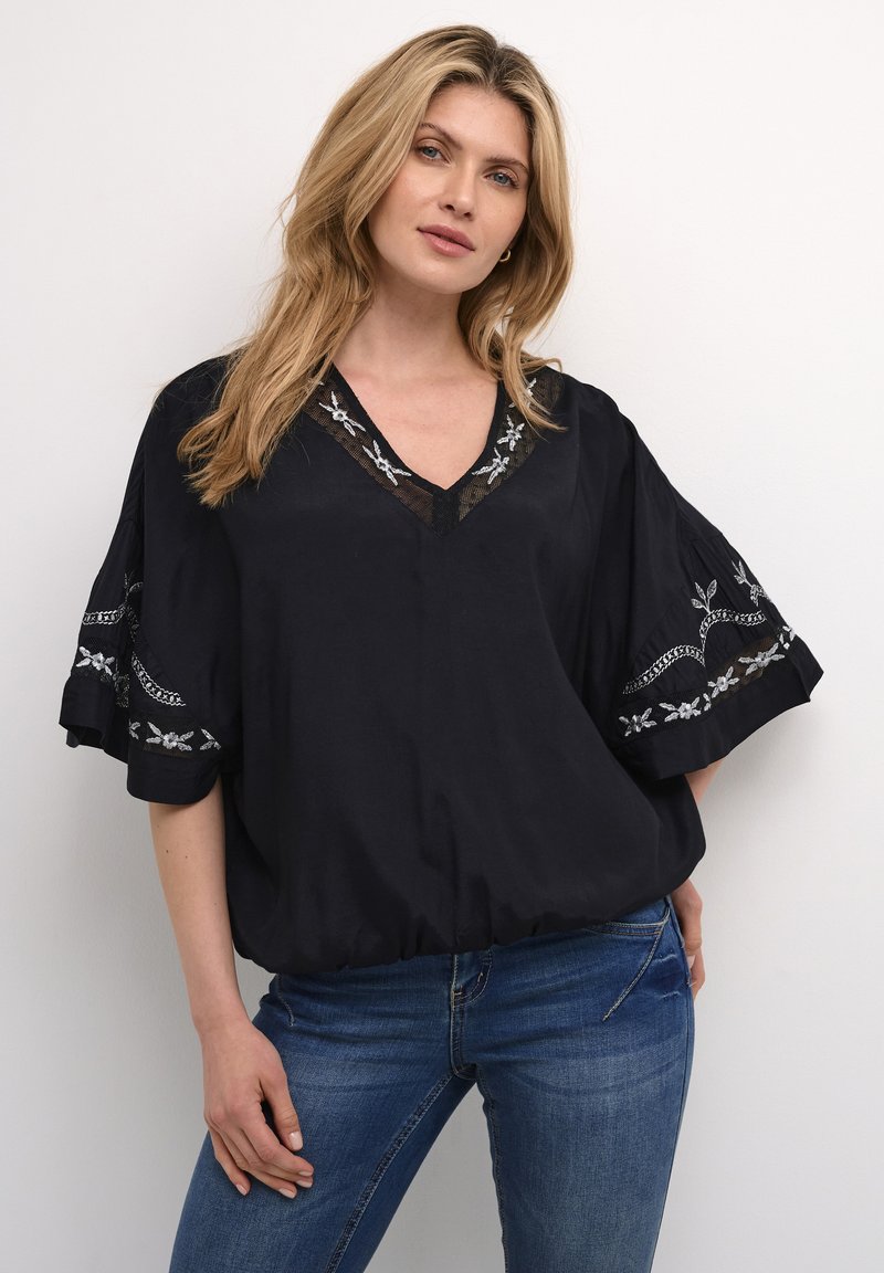Blusa nera con scollatura a V e accenti floreali ricamati sulle maniche. Design morbido e fluido, texture delicata e fondo arricciato. Indossata con jeans blu.