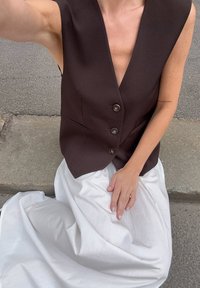 Rjav brezrokavnik z globokim V-izrezom in tremi gumbi, v paru z belim, tekočim krilom. Gladke teksture tkanine in minimalne gubice.