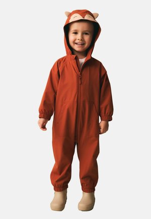 Lächendes Kind trägt einen rostfarbenen Kapuzen-Overall mit Tiergesicht und Ohren auf der Kapuze, beigefarbene Stiefel, steht vor einfachem Hintergrund.