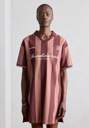 Άτομο που φοράει ένα φαρδιά, κάθετα ριγέ μαρουό και ροζ φόρεμα ποδοσφαίρου με το κείμενο "Scandinavian Football Community" μπροστά.