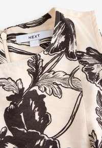 Tissu beige à imprimé floral noir, gros plan montrant le col, la fermeture éclair et une étiquette blanche avec le nom de la marque « NEXT ».