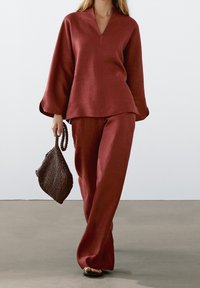 Haut en lin couleur rouille avec un col en V et des manches larges, accompagné d'un pantalon à jambes larges assorti. Le mannequin porte un sac à main tissé marron.