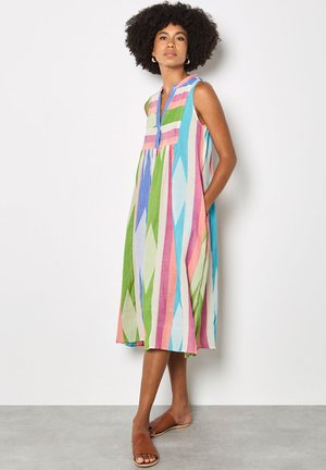 Kobieta z afro irokezem, ubrana w sukienkę bez rękawów o długości do kolan, zdobioną pionowymi kolorowymi geometrycznymi paskami, oraz brązowe sandały wsuwane.