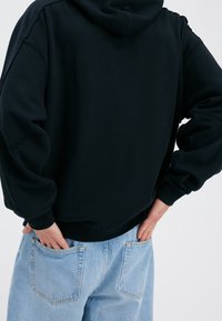 Sudadera negra con capucha, corte relajado, puños acanalados y un acabado texturizado, combinada con unos vaqueros de mezclilla azul claro que cuentan con bolsillos traseros.