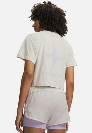 Vrouw met krullend haar, gekleed in een lichtgrijs cropped top met korte mouwen en bijpassende shorts over lila strakke shorts, van achteren gezien.