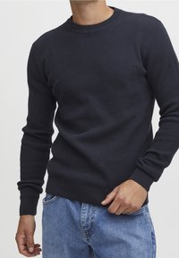 Homme portant un pull à manches longues bleu marine texturé et un jean bleu clair, main près de la poche, torse et bras visibles.