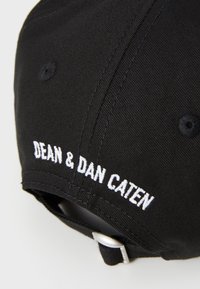 Gorra de béisbol negra de tela con texto bordado en blanco "DEAN & DAN CATEN" en la parte trasera, con un broche de metal para ajuste.