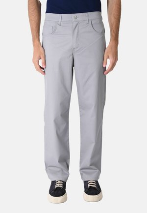Uomo con pantaloni grigio chiaro a gamba dritta e scarpe da ginnastica nere con suole e lacci bianchi, in piedi davanti a uno sfondo semplice.