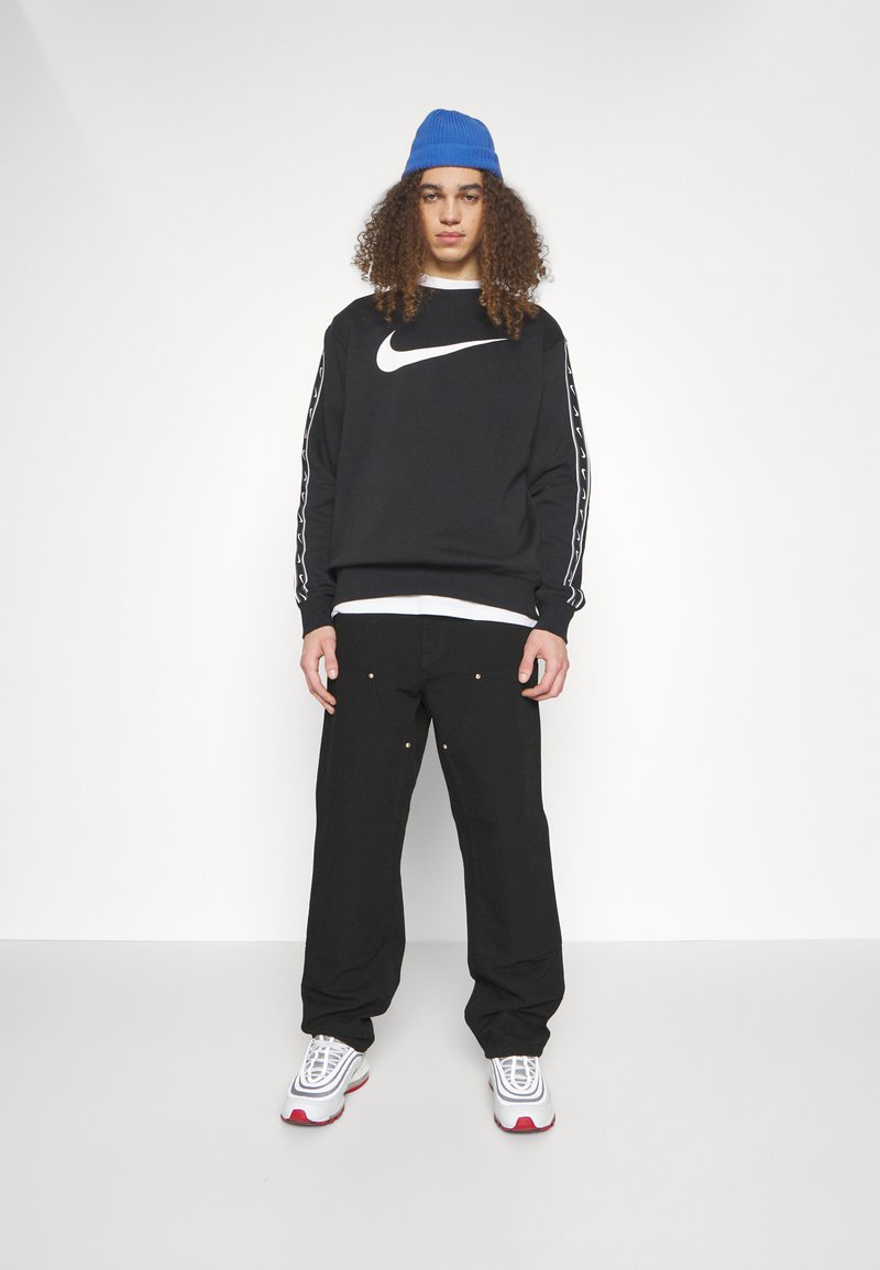 Zwarte Nike sweatshirt met wit logo en zijstrepen, gecombineerd met losse zwarte broek met gouden accenten. Blauwe gebreide muts en witte sneakers.