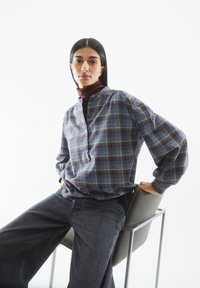 Blau- und graublau-karierter Hemd mit hohem Kragen und Knopfverschluss, kombiniert mit dunkelgrauen, weit geschnittenen Jeans. Das Model sitzt auf einem Stuhl.
