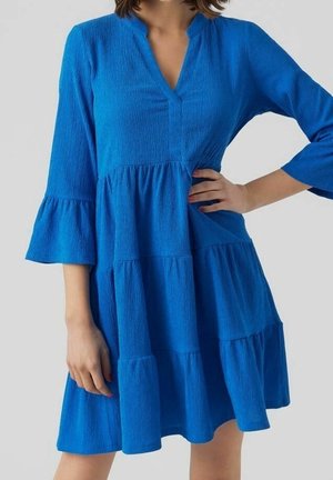 Woman wearing a bright blue, textured tiered dress with 3/4 bell sleeves and a small V-neckline, posing with one hand on her hip.