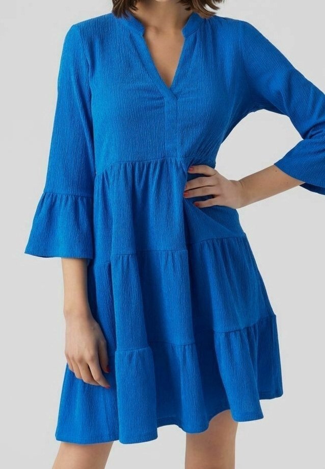 Femme portant une robe à volants texturée bleu vif avec des manches cloche aux trois quarts et un petit décolleté en V, posant avec une main sur la hanche.