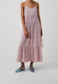 Kwiatowa sukienka maxi w różowym kolorze z cienkimi ramiączkami, marszczonym biustem i falbaną na dole. W zestawieniu z niebieskimi płaskimi butami i małą różową torebką.