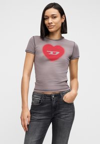 Γυναίκα με ίσια σκούρα μαλλιά, φορώντας ένα εφαρμοστό γκρι μπλουζάκι με κόκκινη καρδιά και λογότυπο, συνδυασμένο με ξεθωριασμένο μαύρο τζιν, που στέκεται με το ένα χέρι στην τσέπη.