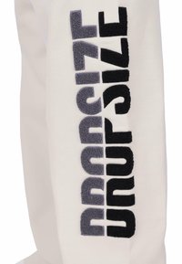 Baltas sporta bikses ar vertikālu tekstūru, kurā ir uzraksts "DROPSIZE" trijās krāsās: melnā, tumši pelēkā un gaiši pelēkā. Mīksta auduma apdare.