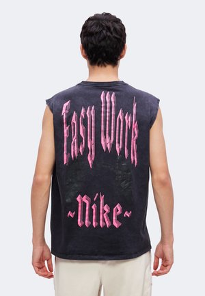 Homme portant un débardeur noir avec un texte rose "Easy Work Nike" dans le dos, debout devant un fond clair uni.