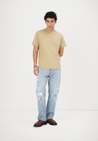 Beige Baumwoll-T-Shirt mit kurzen Ärmeln und Rundhalsausschnitt. Hellblaue Jeans mit Rissen. Braune Leder Schuhe mit niedrigem Profil. Schlicht und lässig.