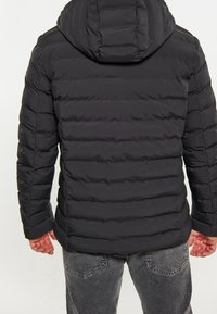 Homme portant une doudoune noire avec capuche et un jean noir délavé, vu de dos sur un fond clair uni.