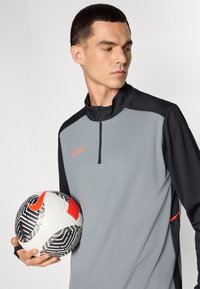 Grijs en zwart sportjack met rits en oranje accenten. Het model houdt een gestreepte zwart-wit voetbal met een rood logo vast.