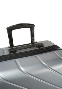 Maleta gris de carcasa dura con ranuras verticales, que cuenta con un asa telescópica negra y un cierre de cremallera. El logo "DKNY" es visible.