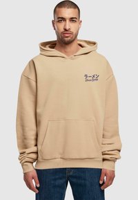 Sudadera beige con capucha que presenta un bolsillo canguro en la parte delantera, un corte holgado y texto impreso en azul oscuro en el área del lado izquierdo del pecho.