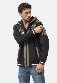 Zwarte capuchonvest met beige accenten, ritssluiting aan de mouwen en wit/rood gestreepte manchetten, voorzien van grafische tekst op de borst en zakken.