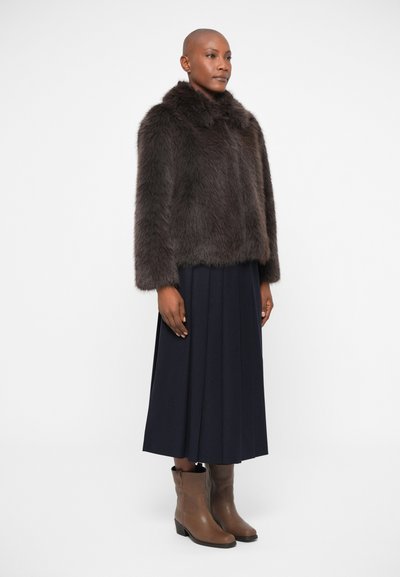 Casaco de pelo sintético castanho com um colar alto, usado sobre uma saia longa plissada azul marinho. Botas de tornozelo castanhas com salto completam o visual.