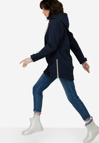 Chaqueta impermeable navy con capucha, bolsillos laterales con cremallera, combinada con botas de tobillo reflectantes ligeras y vaqueros azules, en pose de caminar.