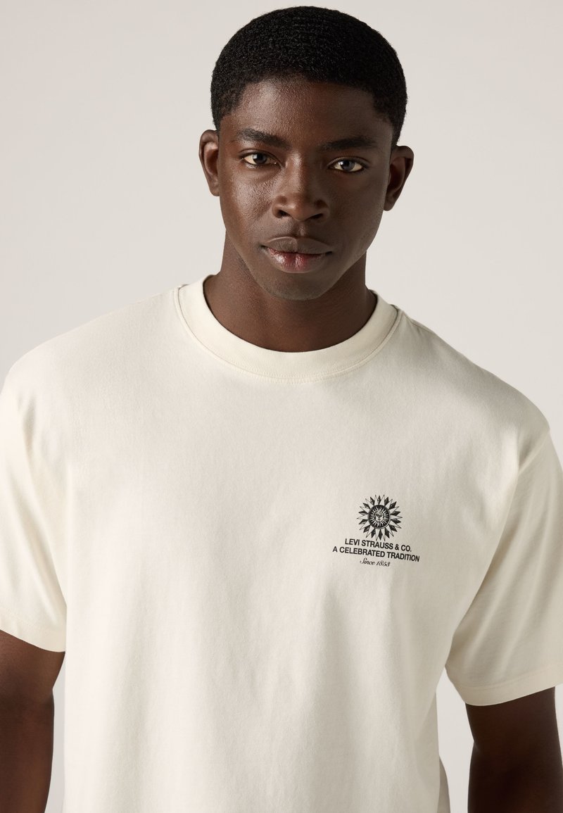 Young man with short hair wears a white Levi Strauss t-shirt with a small black logo and text on the left chest.