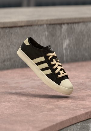 Μαύρο και μπεζ αθλητικό παπούτσι Adidas με τρεις ρίγες και κορδόνια, κρεμασμένο πάνω από μια πέτρινη επιφάνεια με θολό γκρι φόντο.
