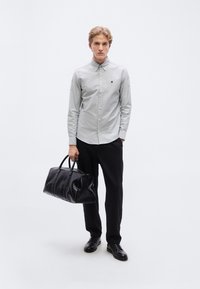 Γκρι πουκάμισο με κουμπιά και μικρό λογότυπο, μαύρη υφασμάτινη τσάντα ταξιδίου, μαύρο παντελόνι και παπούτσια, σε απλό λευκό φόντο.