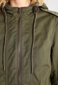 Giacca leggera verde oliva con zip frontale, lacci per cappuccio regolabili e texture sottile. Presenta epaulettes sulle spalle e hardware in bronzo.