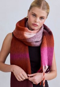 Zachte, gradient sjaal met warme tinten van rood, roze en paars. Het materiaal is fuzzy, met een textuurafwerking en franjes aan de randen.