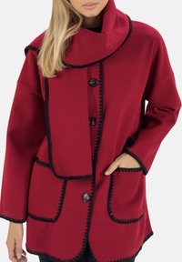 Chaqueta roja con cuello alto, ribete negro, cierres de botones y dos bolsillos delanteros. Confeccionada con tela texturizada y ajuste holgado.