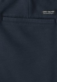 Tissu texturé bleu foncé avec une poche horizontale fendue et une étiquette noire indiquant "STREET ONE MEN Unmatched Comfort."