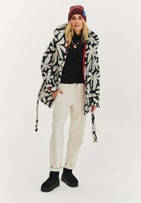 Černobílý kabát s květinovým vzorem, červený vnitřek, doplněný černým topem, krémovými kalhotami a černými krátkými kotníkovými botami. Součástí je také vínová čepice.