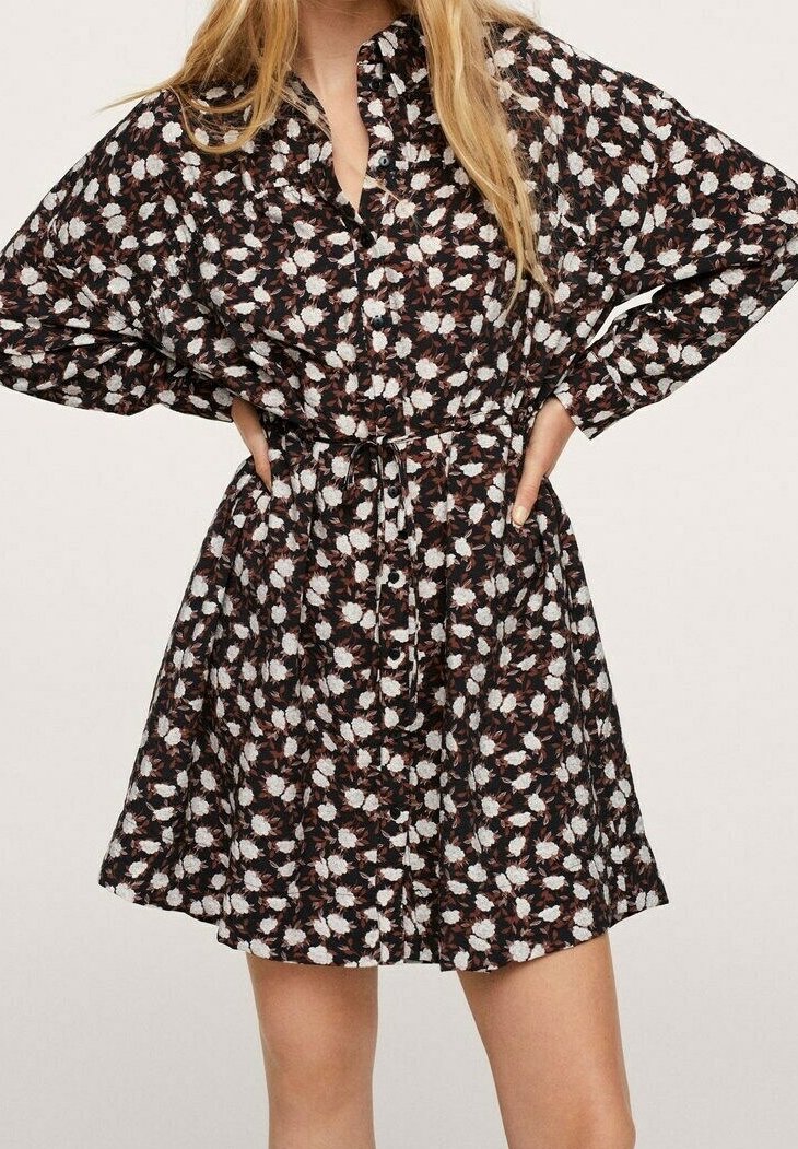 Femme portant une robe chemise noire à motif floral, manches longues et ceinture à la taille, debout les mains sur les hanches.
