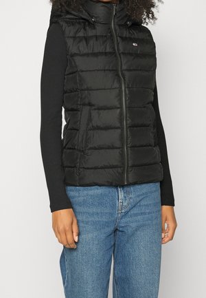 Osoba ubrana w czarną pikowaną kamizelkę bez rękawów na czarnym, długim rękawie i niebieskich dżinsach, stojąca na tle jasnego, jednolitego tła.