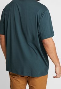 Donker tealkleurig katoenen T-shirt met korte mouwen, ontspannen pasvorm en rechte zoom. Draagt met beige broek, toont eenvoudige achteraanzicht.